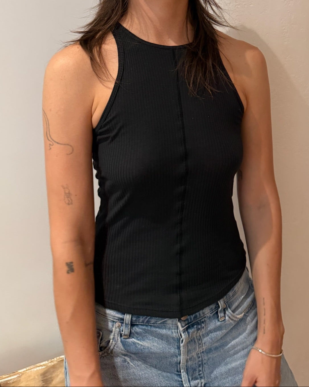 A woman with dark hair wearing a black ribbed sleeveless halter-style top and light blue jeans, standing against a plain beige wall.