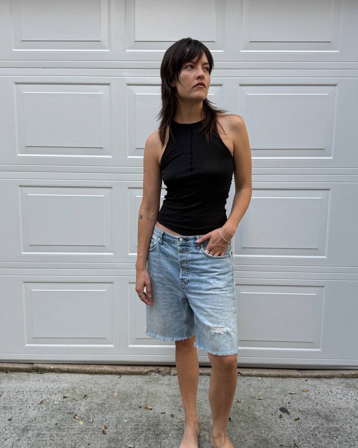 A woman stands barefoot in front of a white garage door, wearing a black sleeveless top and light-wash denim shorts.
