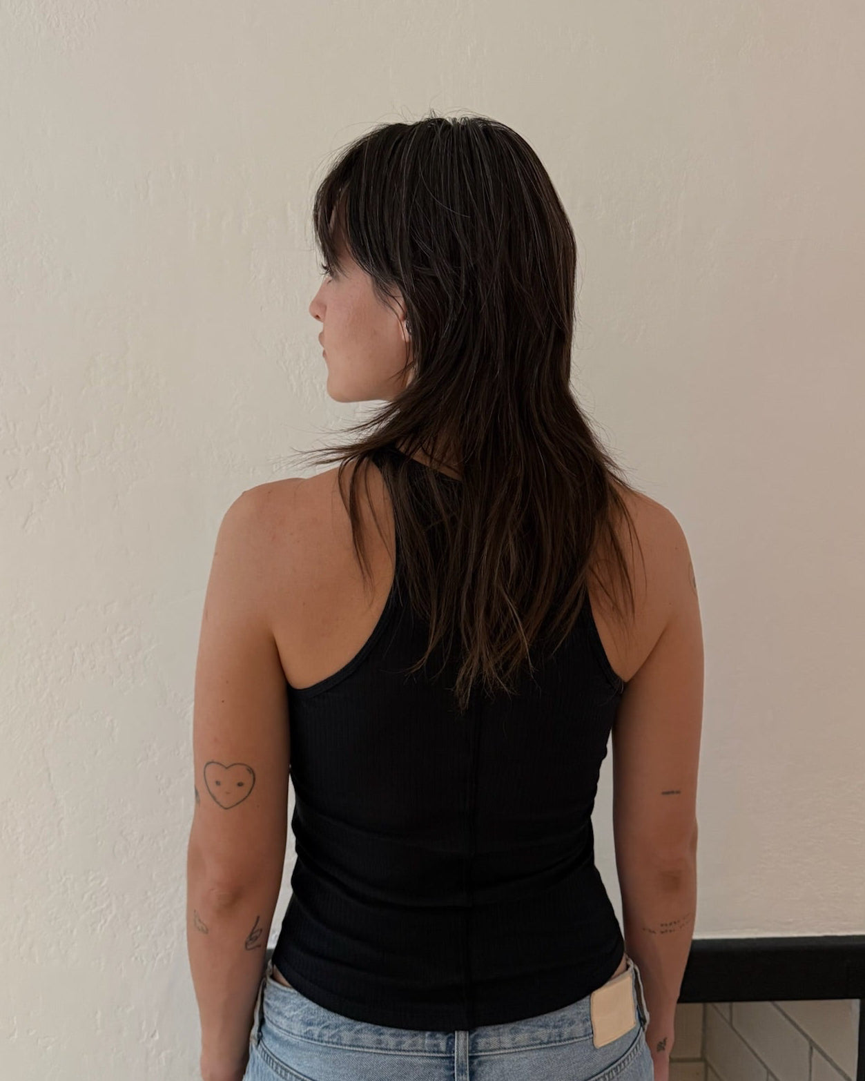 A woman with long dark hair is shown from behind wearing a black ribbed tank top and light blue jeans, standing against a plain beige wall.